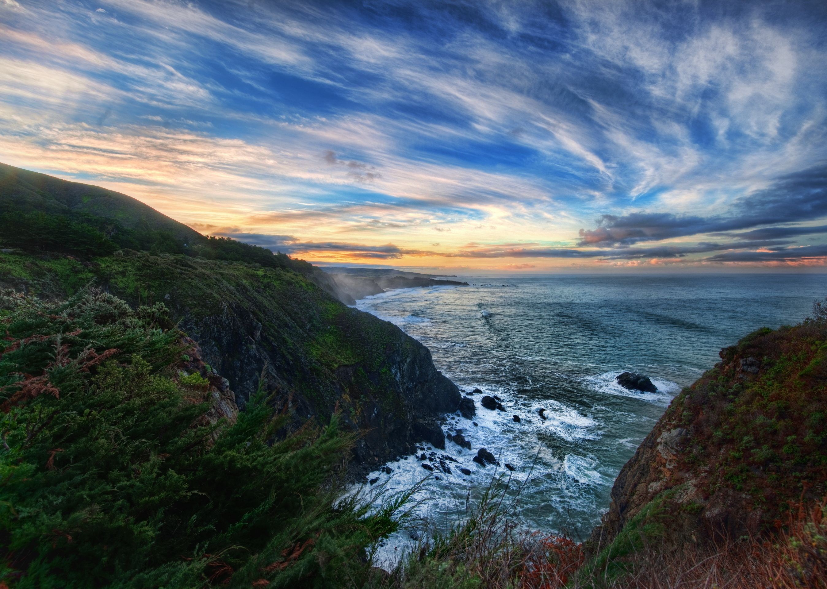 Dramatic sunset over a rugged coastline with crashing waves HD Wallpaper
