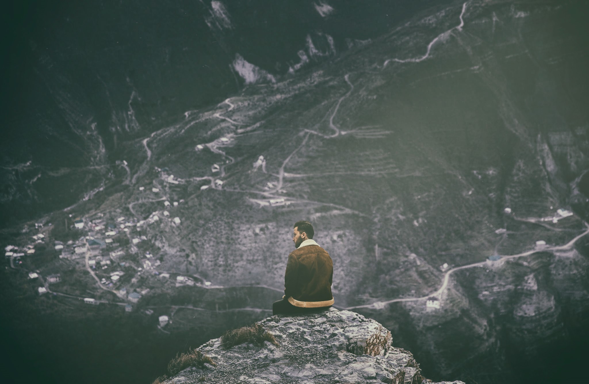 Man sitting on a cliff overlooking a village HD Wallpaper