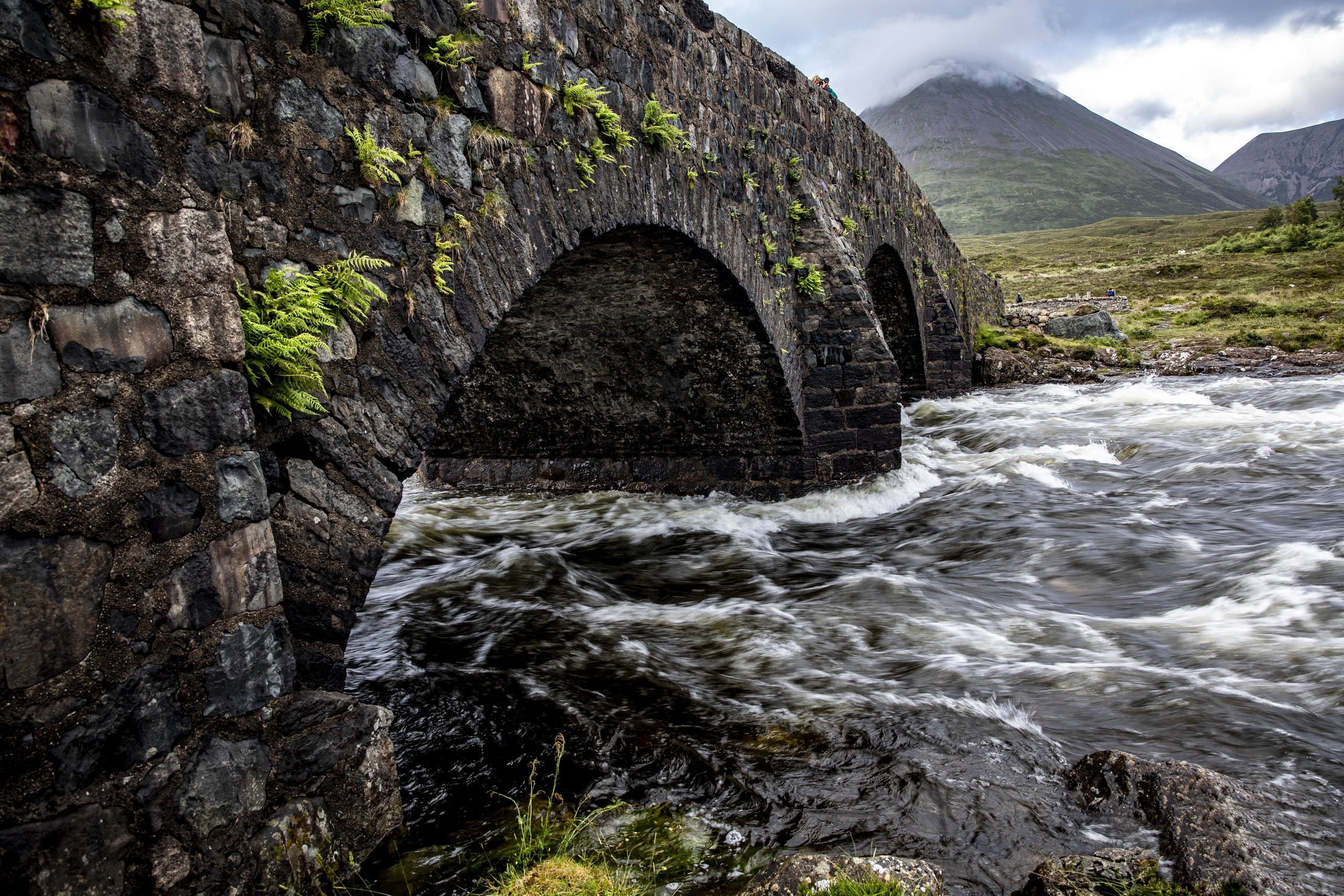 Stone arch bridge over a flowing river with mountains in the background HD Wallpaper
