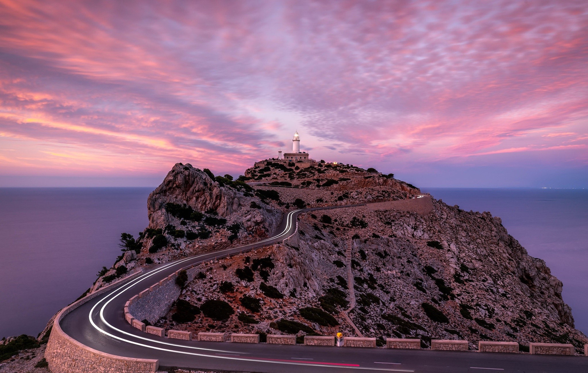 Lighthouse on a rocky cliff with a winding road at sunset HD Wallpaper