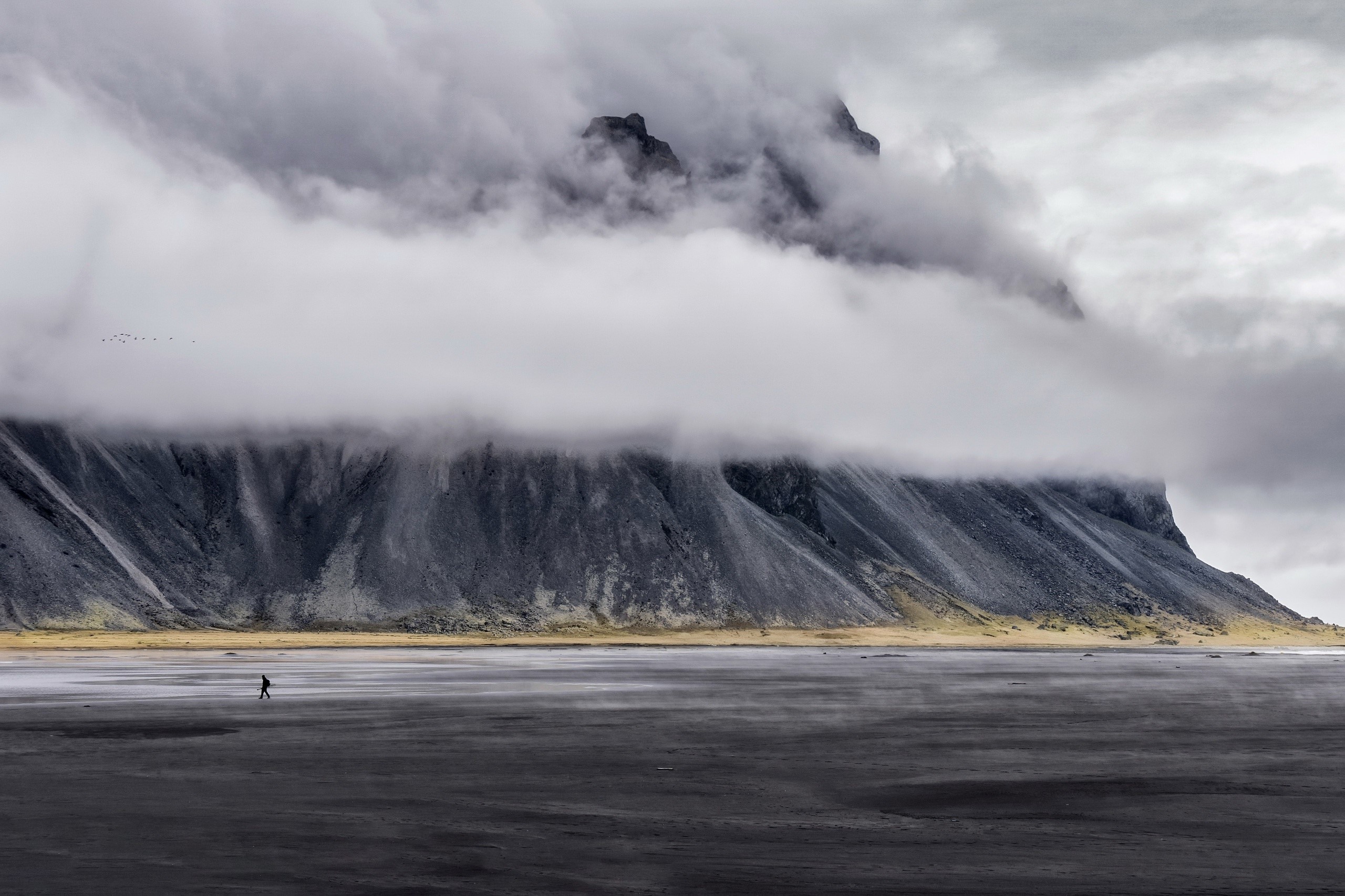 Person walking on a black sand beach with mountains and clouds HD Wallpaper
