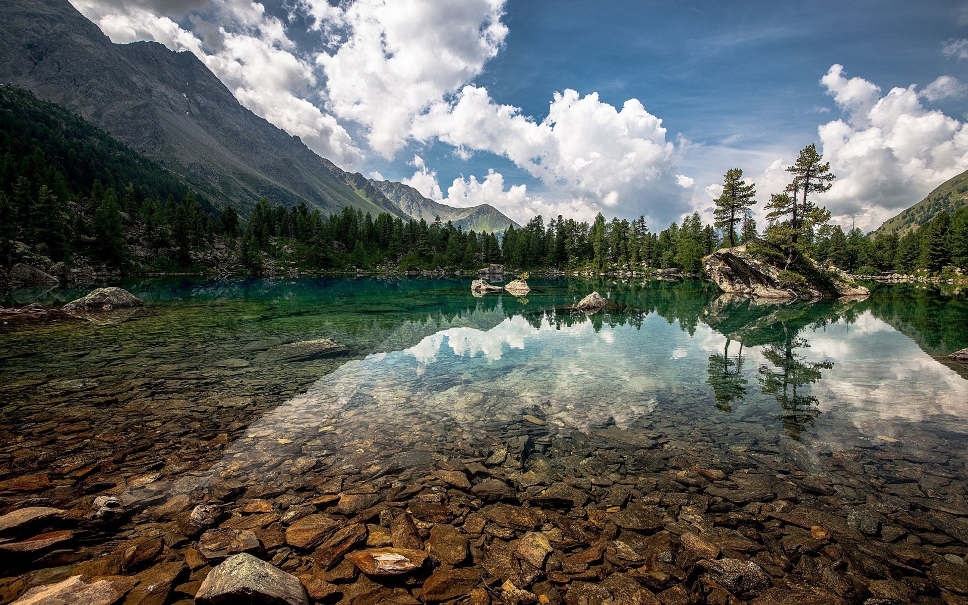 Clear lake reflecting mountains and forest with rocky shore HD Wallpaper