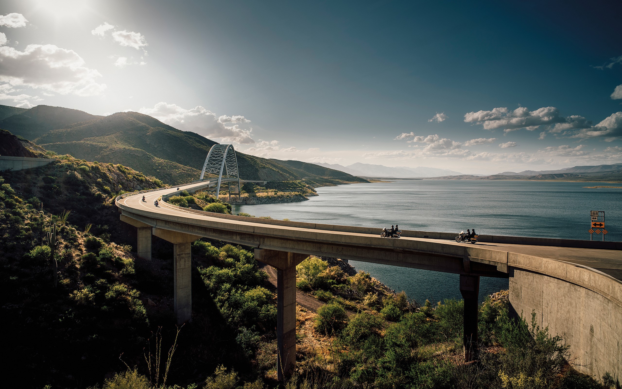 Motorcyclists on a bridge over a lake with mountains HD Wallpaper