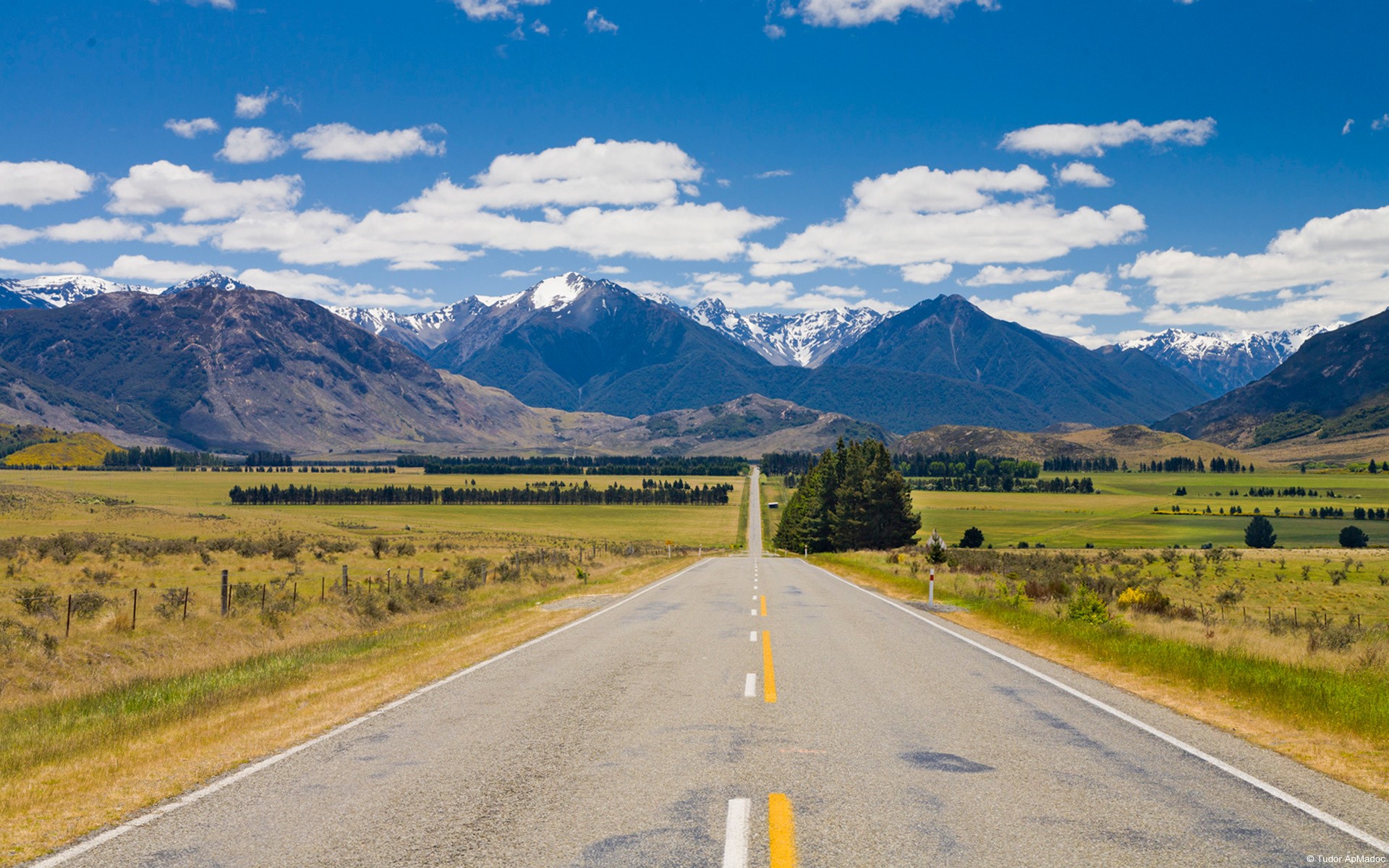 Long road leading to snow-capped mountains under a blue sky HD Wallpaper