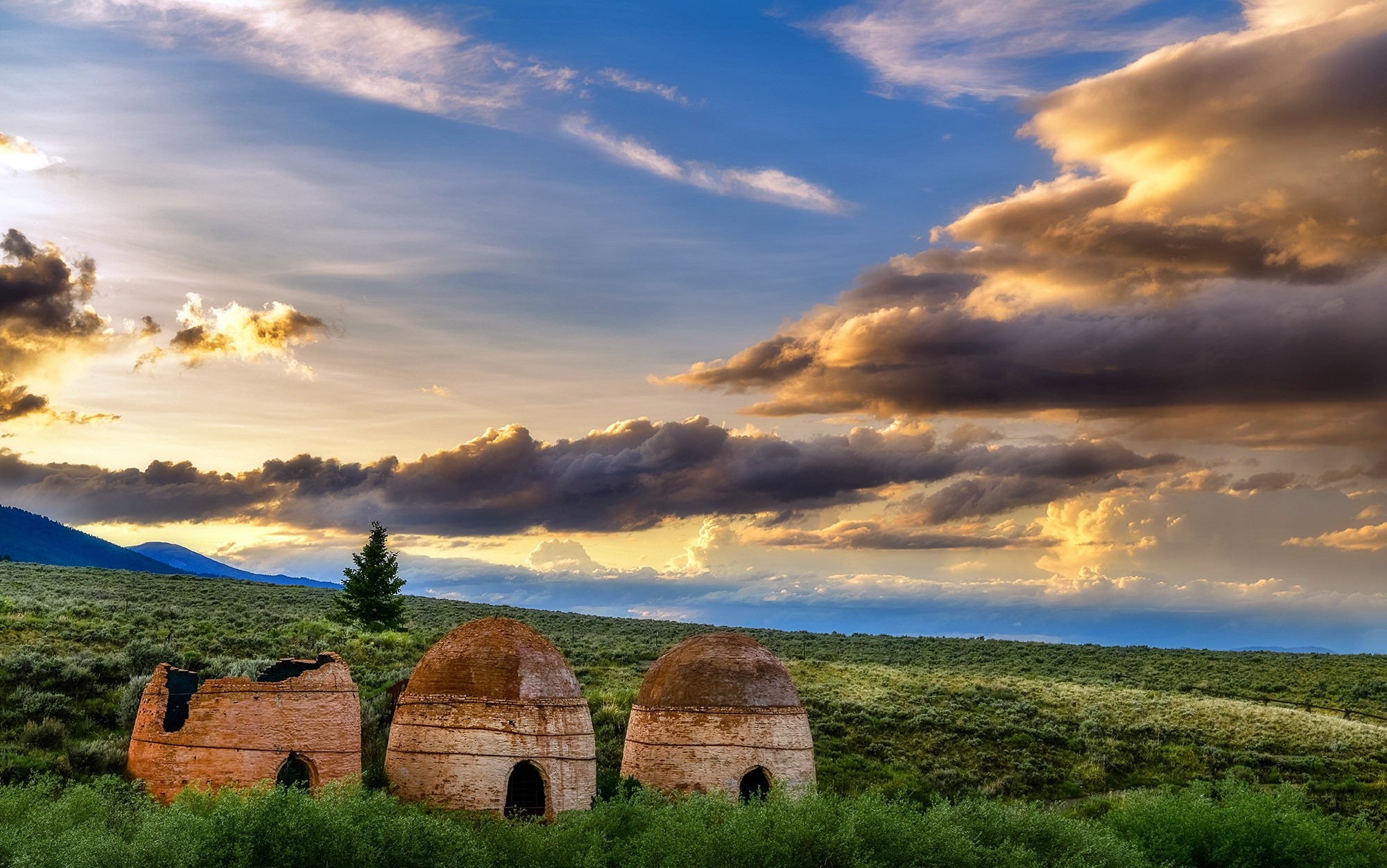 Old kilns in a grassy field under a cloudy sky HD Wallpaper