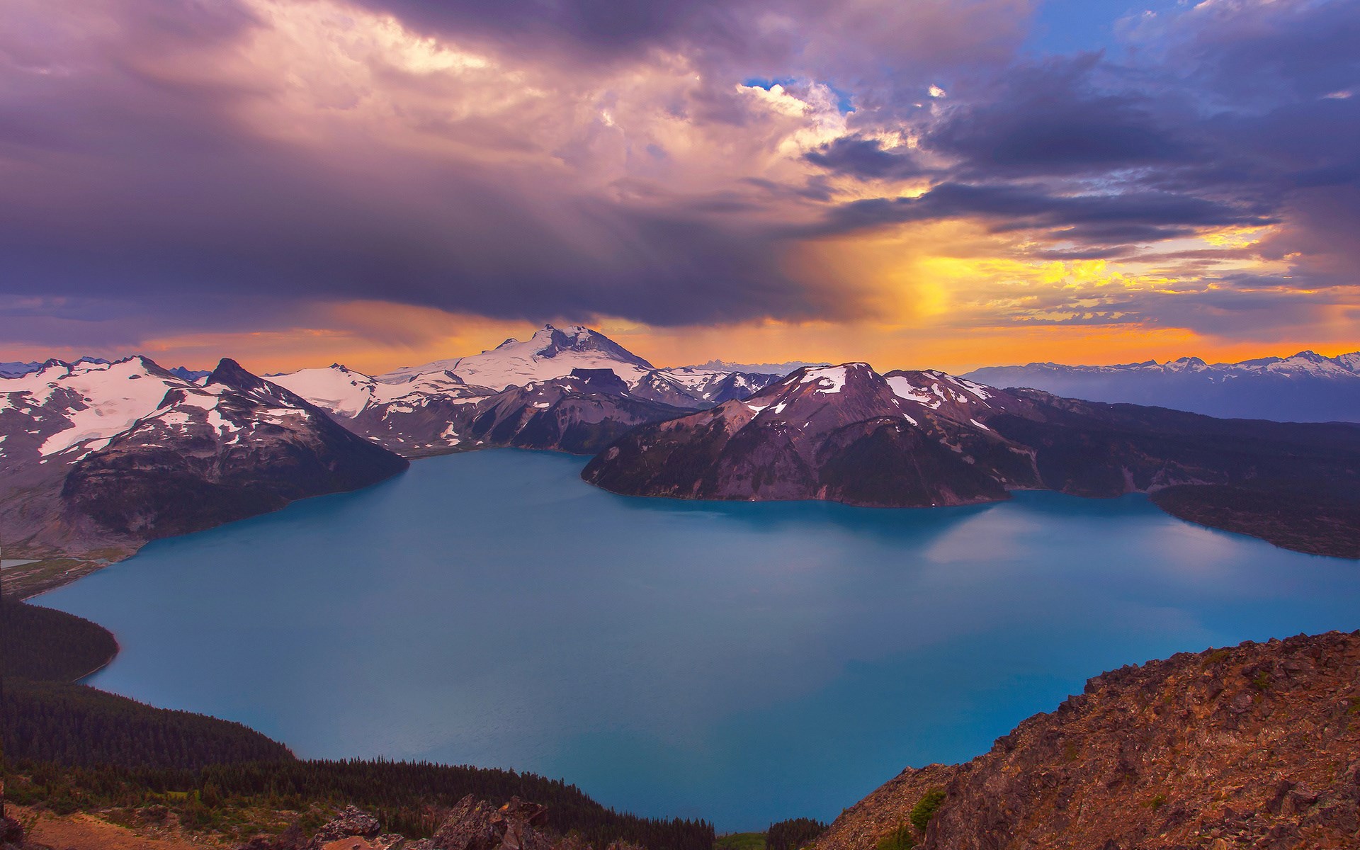Turquoise lake surrounded by snow-capped mountains under a dramatic sky HD Wallpaper