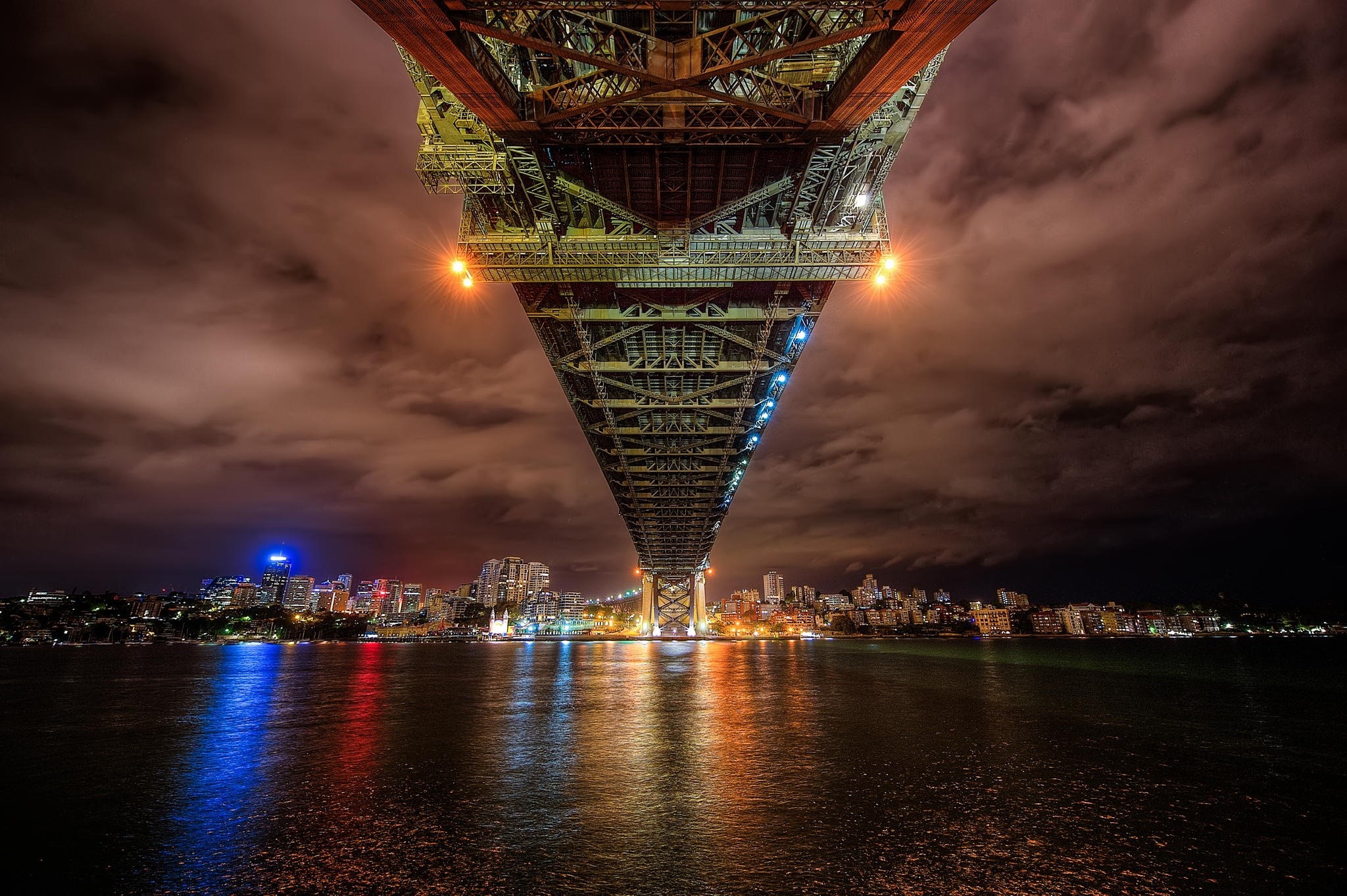 Underneath a bridge at night with city lights reflecting on water HD Wallpaper