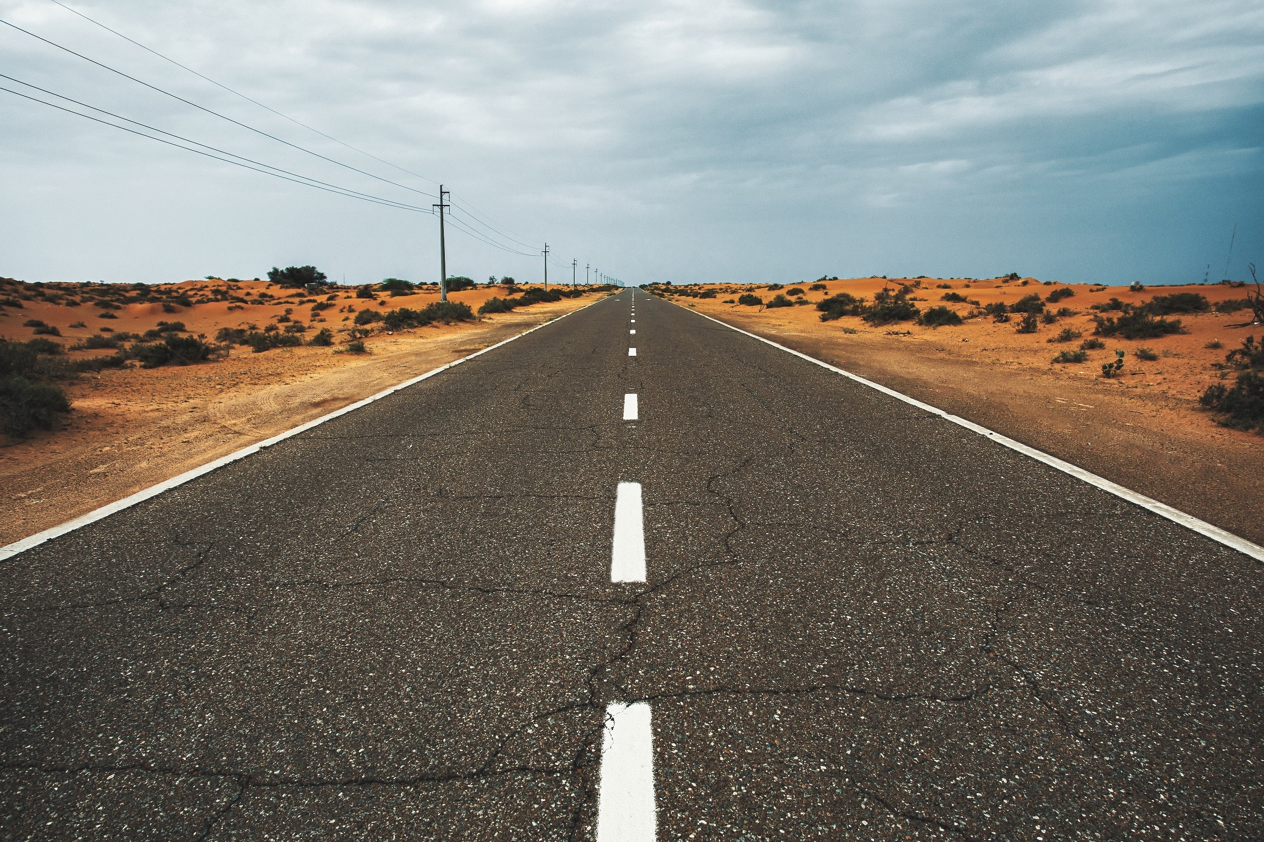 Desert road with power lines under a cloudy sky HD Wallpaper