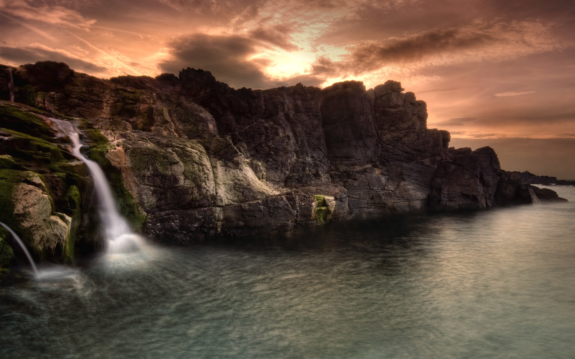 Waterfall cascading over rocks into the sea at sunset HD Wallpaper