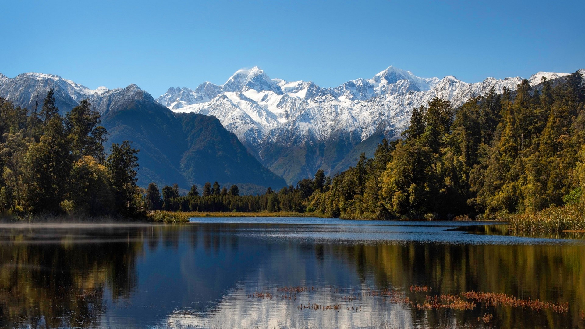 Snow-capped mountains reflected in a calm lake with trees HD Wallpaper