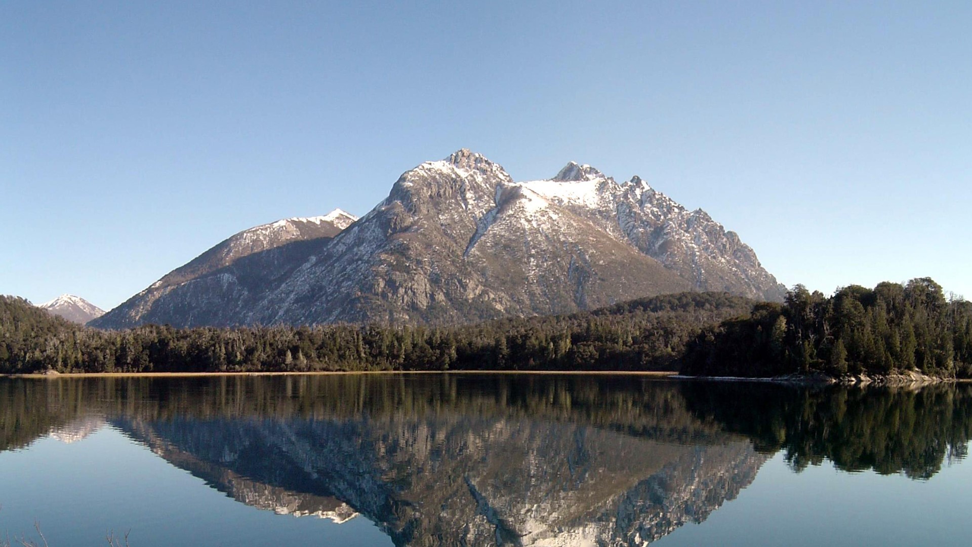 Snow-capped mountains reflected in a lake with pine forest HD Wallpaper
