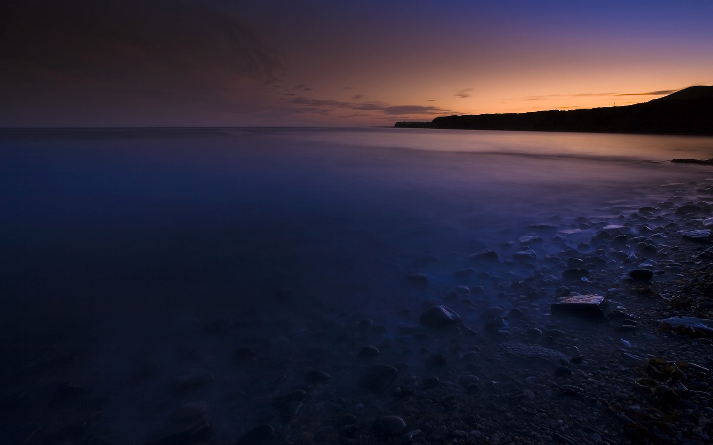 Dark beach at dawn with calm ocean and rocky shore HD Wallpaper