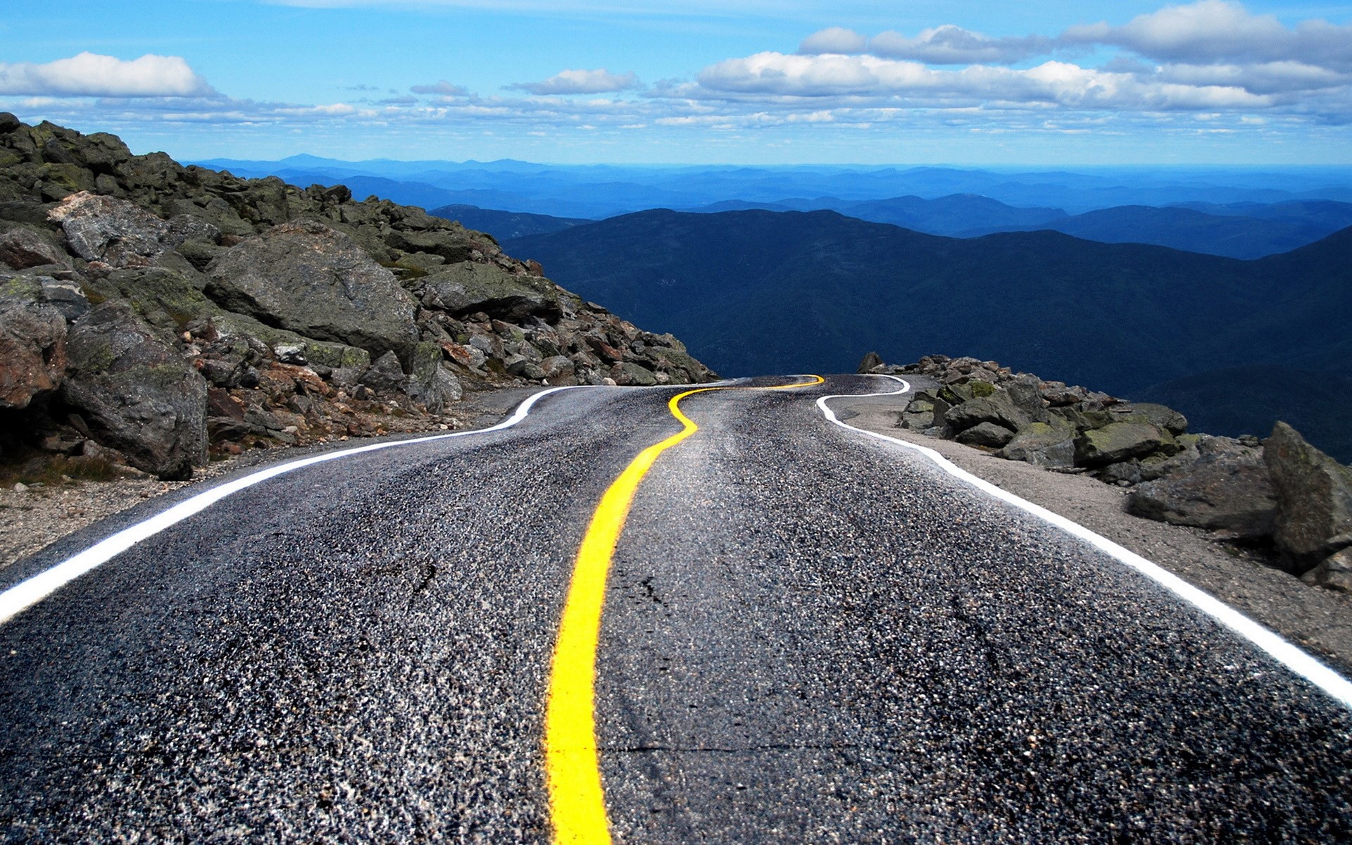 Winding mountain road with rocky terrain and distant blue mountains HD Wallpaper