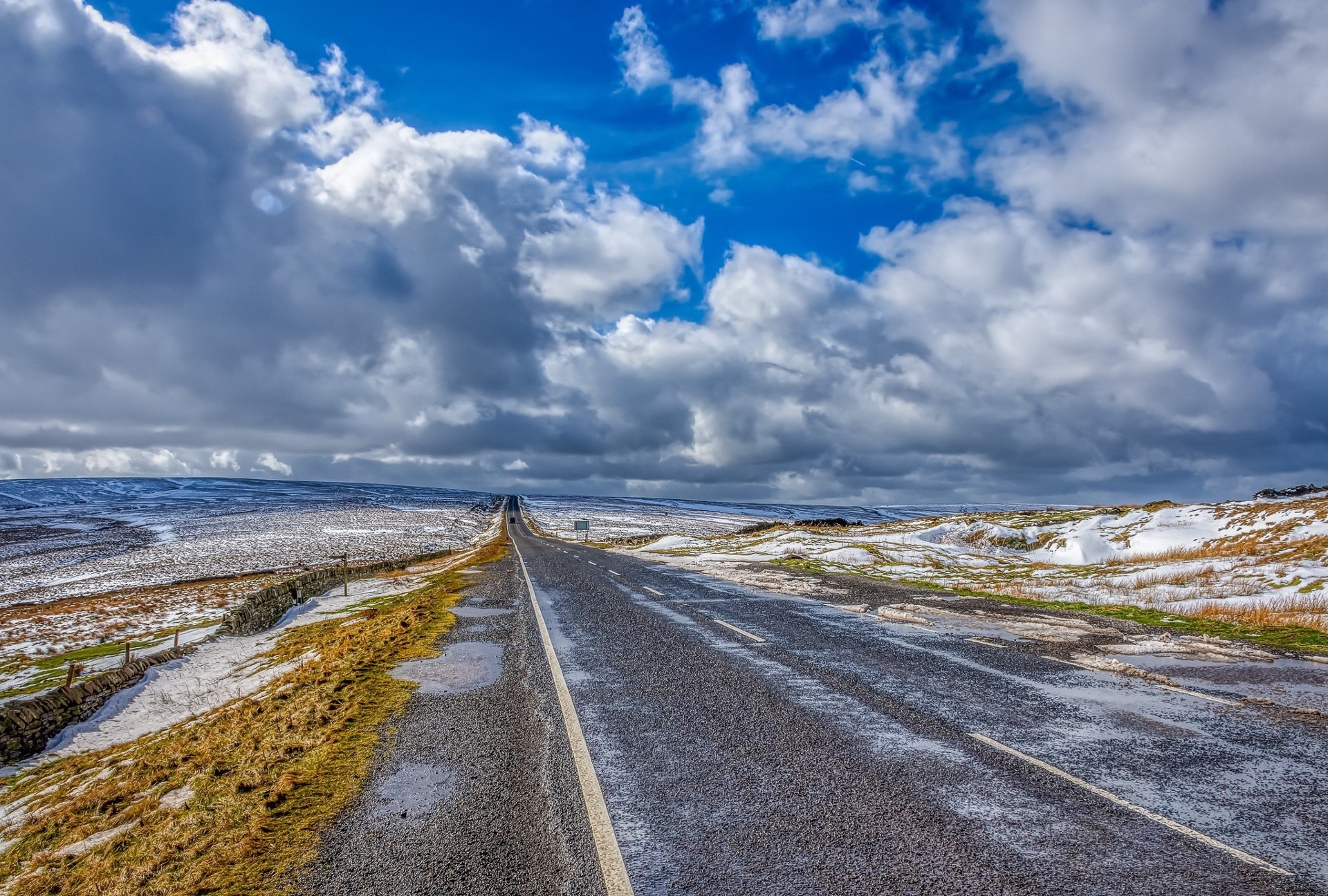 A long, wet asphalt road stretches through a snowy landscape under a cloudy sky. HD Wallpaper