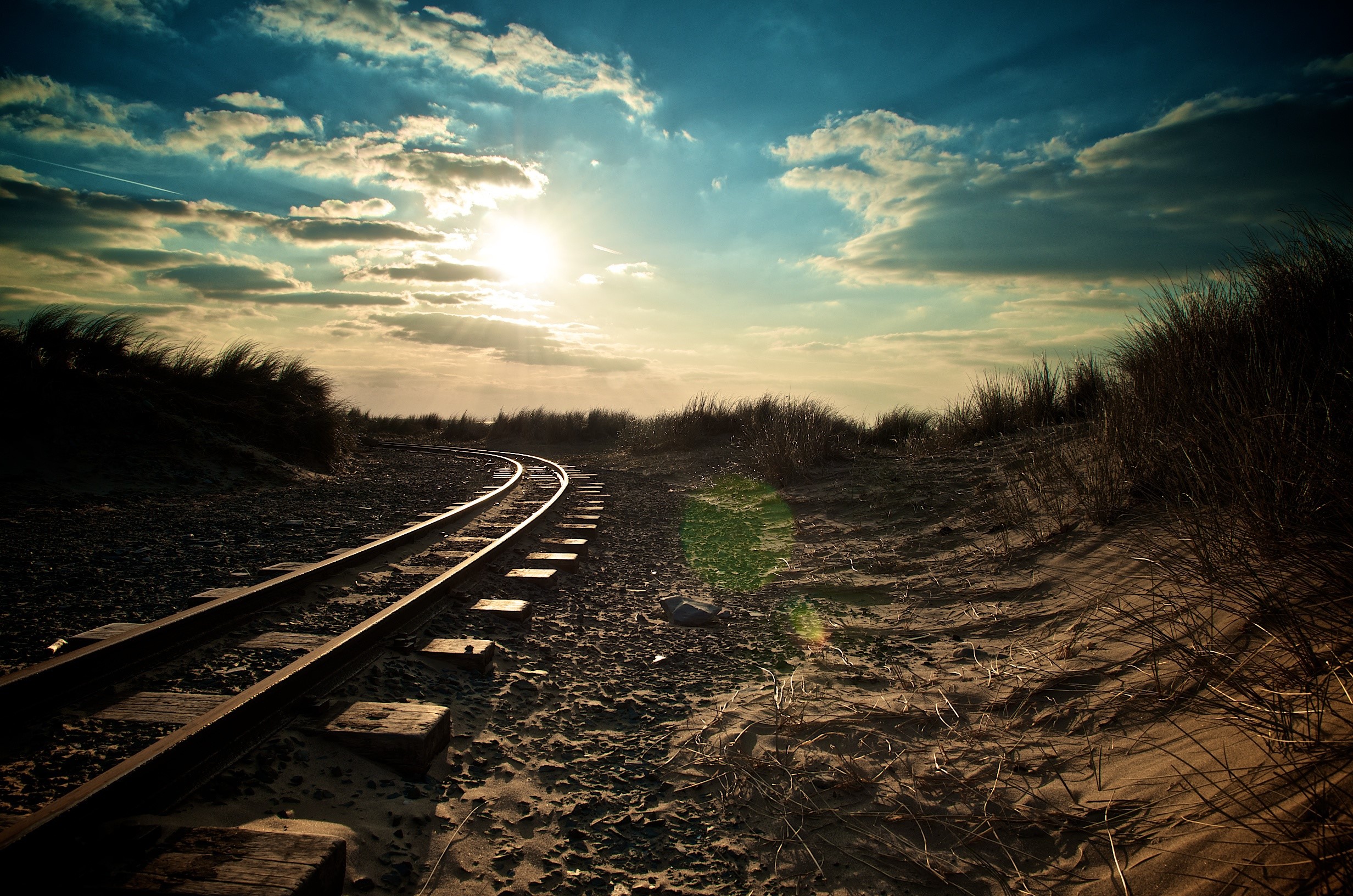 Railway tracks through sand dunes under a cloudy sky HD Wallpaper