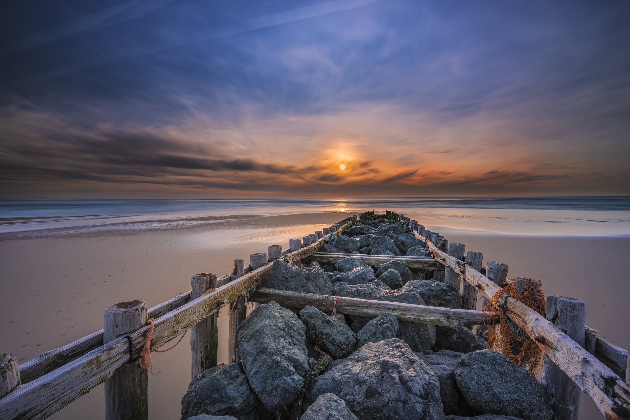 Sunset over a rocky breakwater on a beach HD Wallpaper
