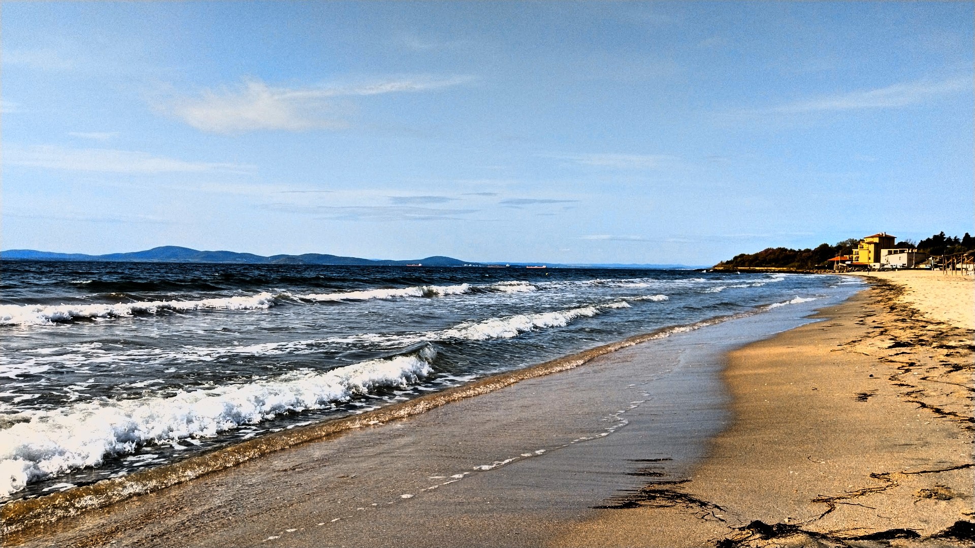 Waves crashing on a sandy beach with buildings in the distance HD Wallpaper