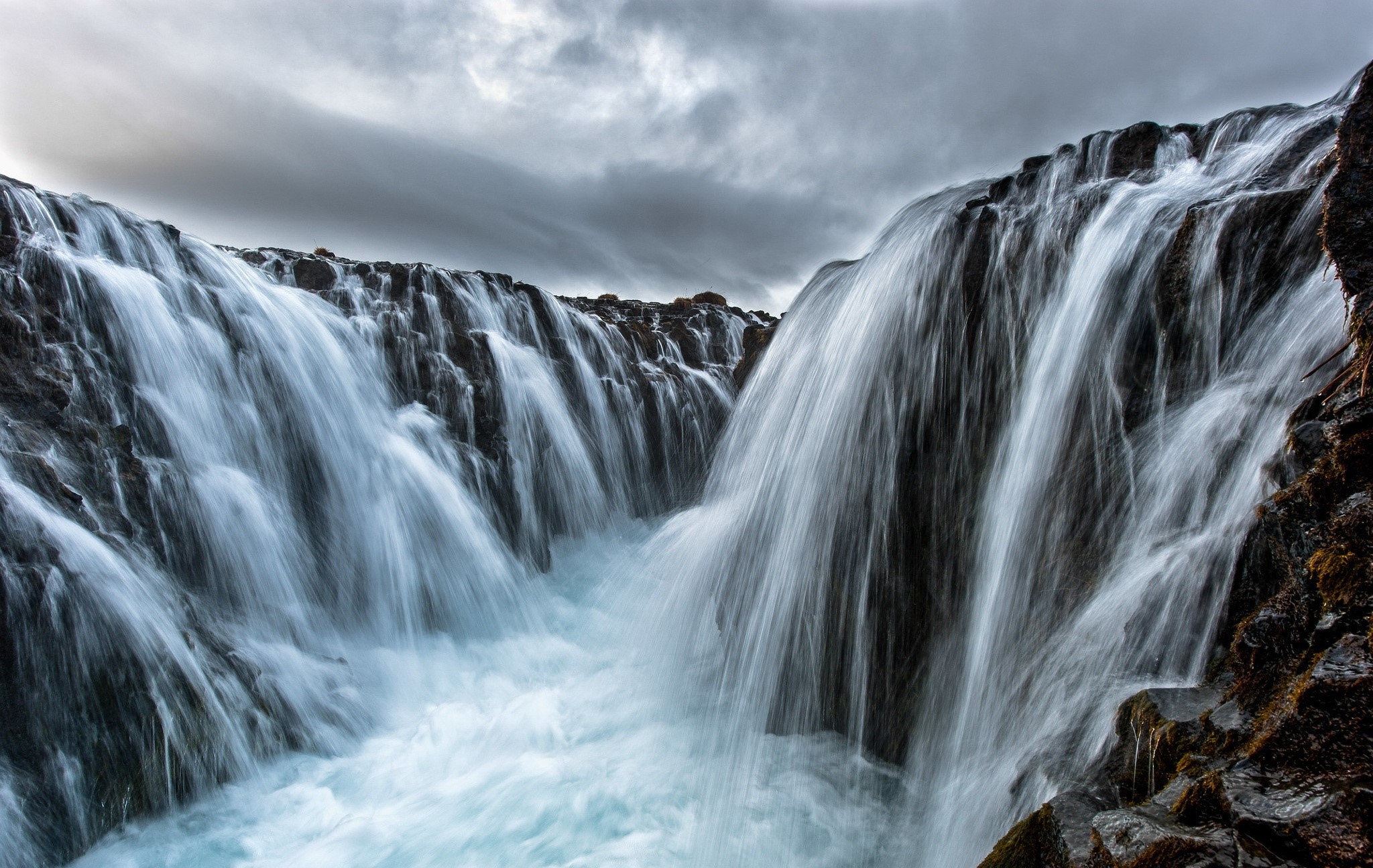 Waterfall cascading over dark rocks under a cloudy sky HD Wallpaper