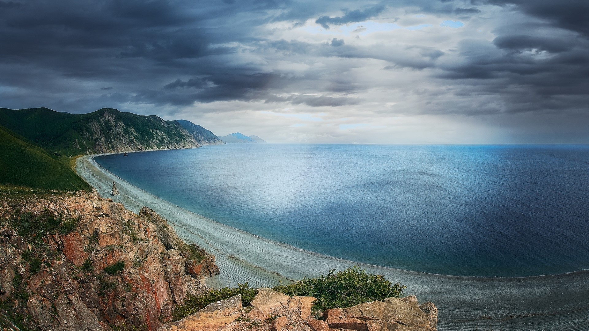 Rocky cliff overlooking a curved beach and the sea under cloudy sky HD Wallpaper