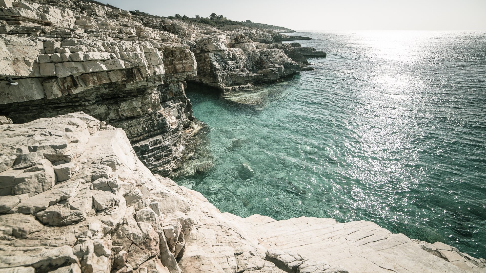 Rocky coast with clear turquoise sea and sunlight reflection HD Wallpaper
