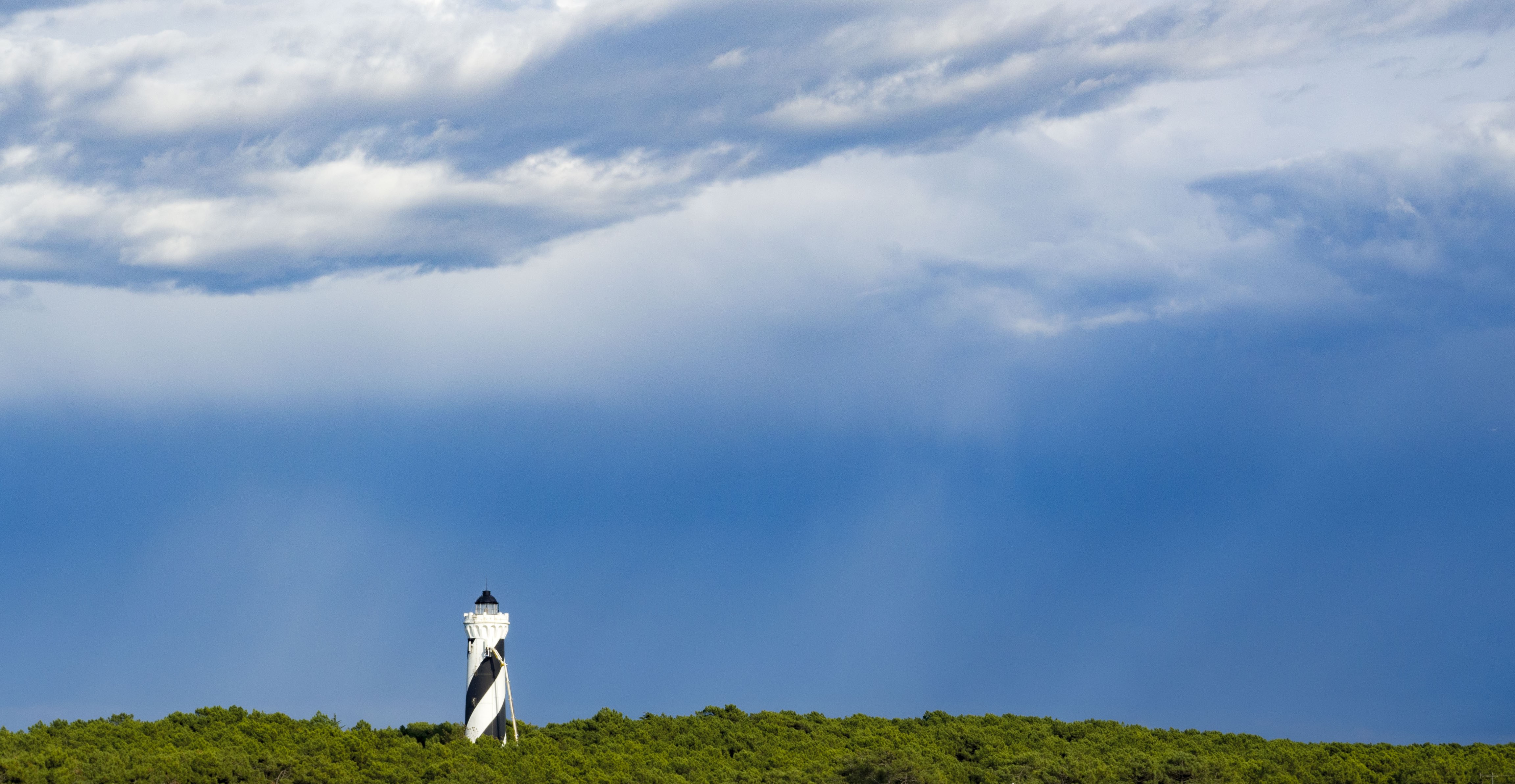 4K Lighthouse on a Tree Line Under Cloudy Sky HD Wallpaper