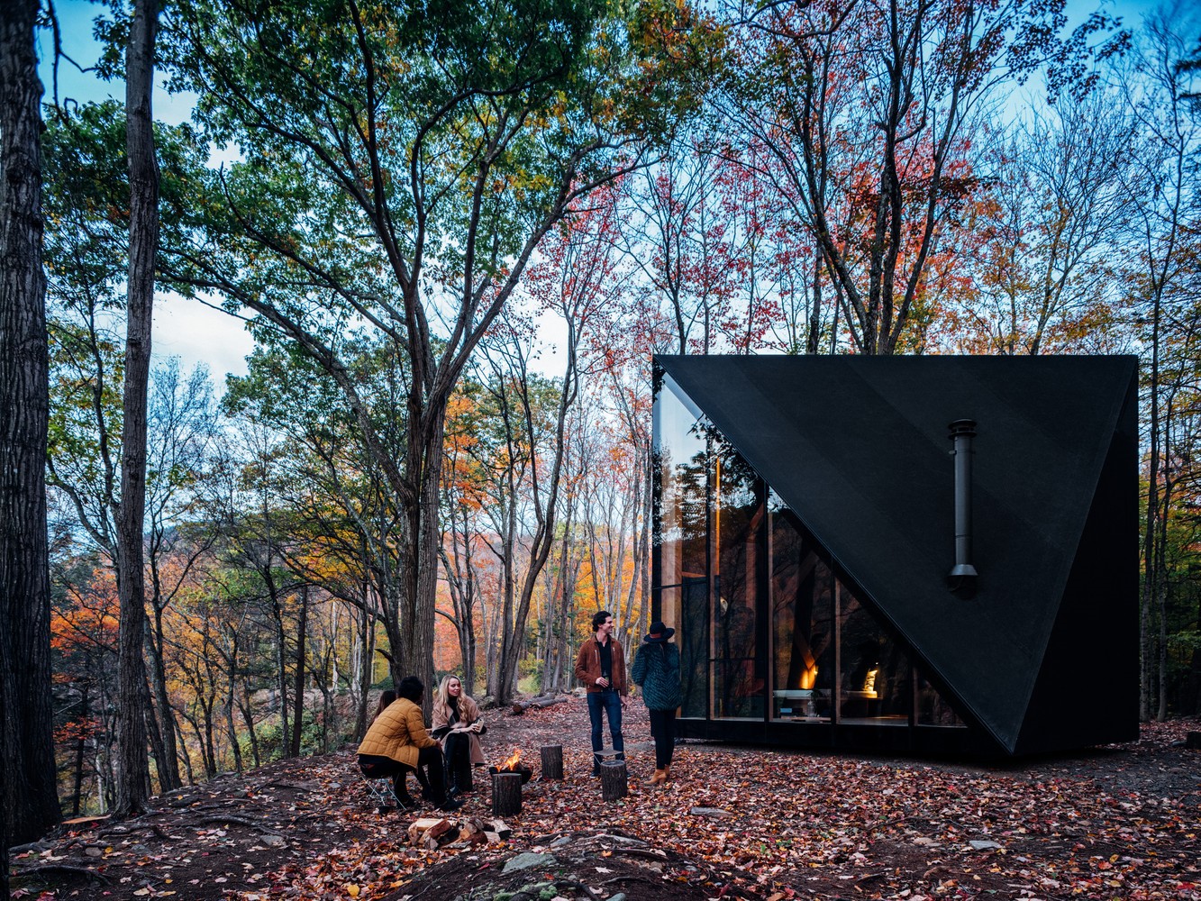 Friends gather around a campfire outside a modern cabin in the woods HD Wallpaper