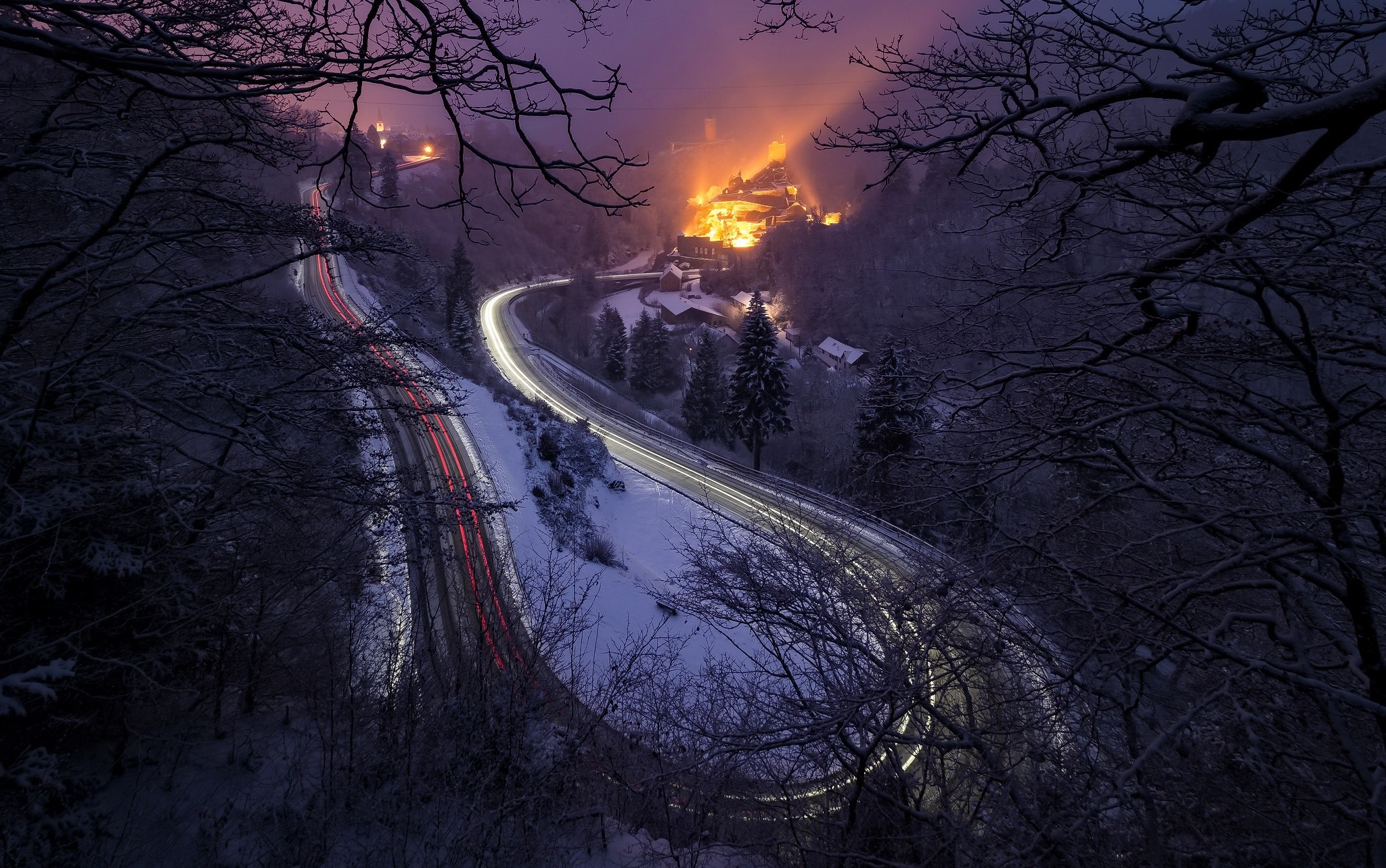 Long exposure of a snowy road with car lights and a village at night HD Wallpaper