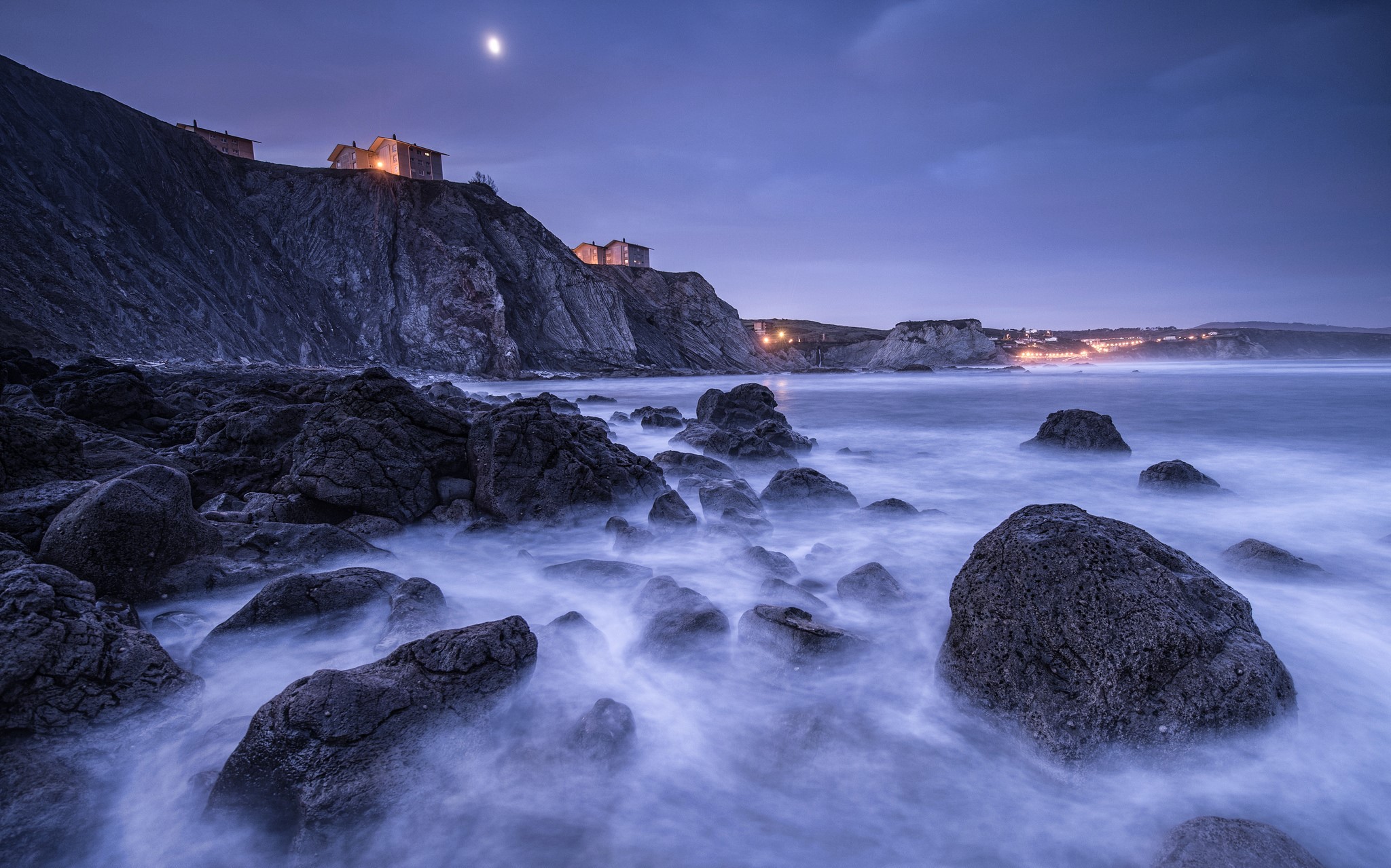 Long exposure of rocky coastline with buildings and moon HD Wallpaper