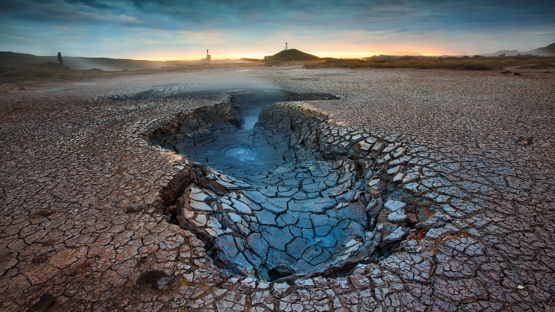 Cracked earth and mud pool landscape with distant structures HD Wallpaper