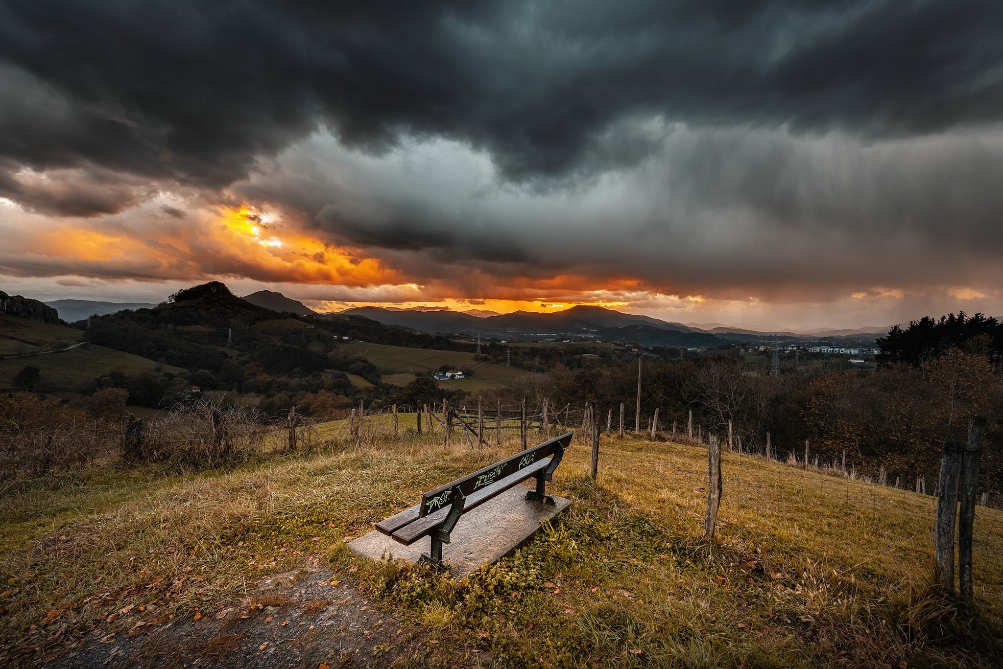 Bench overlooking a stormy sunset landscape HD Wallpaper