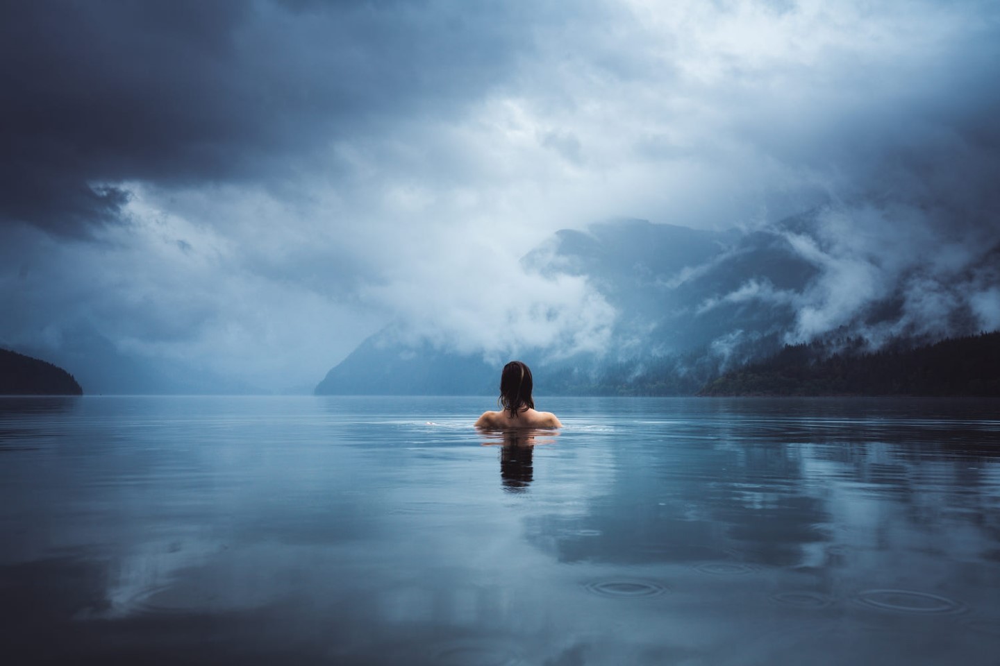 Person swimming in a lake with mountains under overcast sky HD Wallpaper