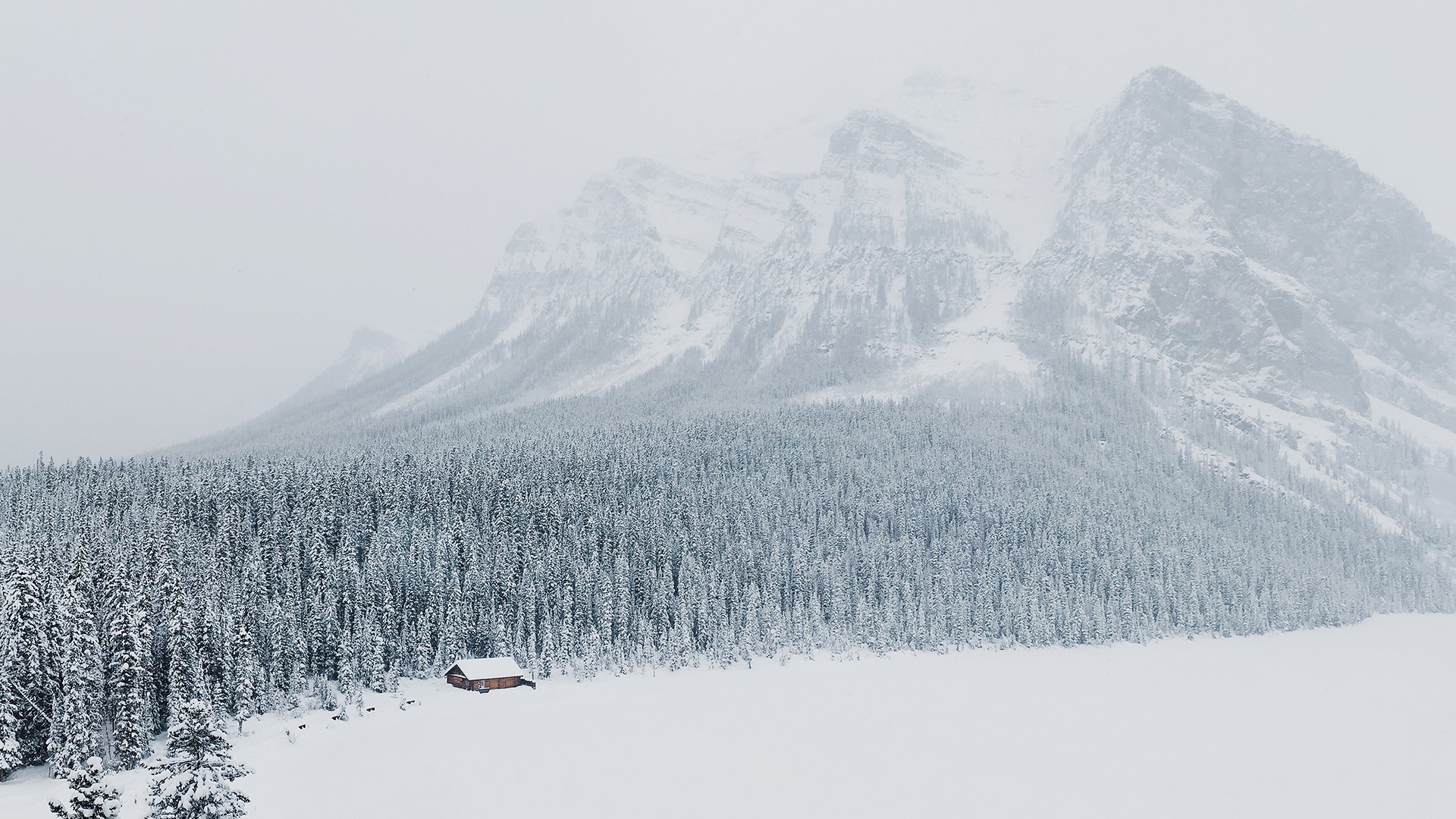Snowy cabin in front of snow-covered mountains and forest HD Wallpaper