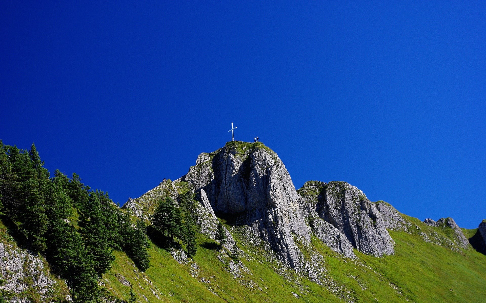 Cross on a rocky mountain peak with green slopes HD Wallpaper