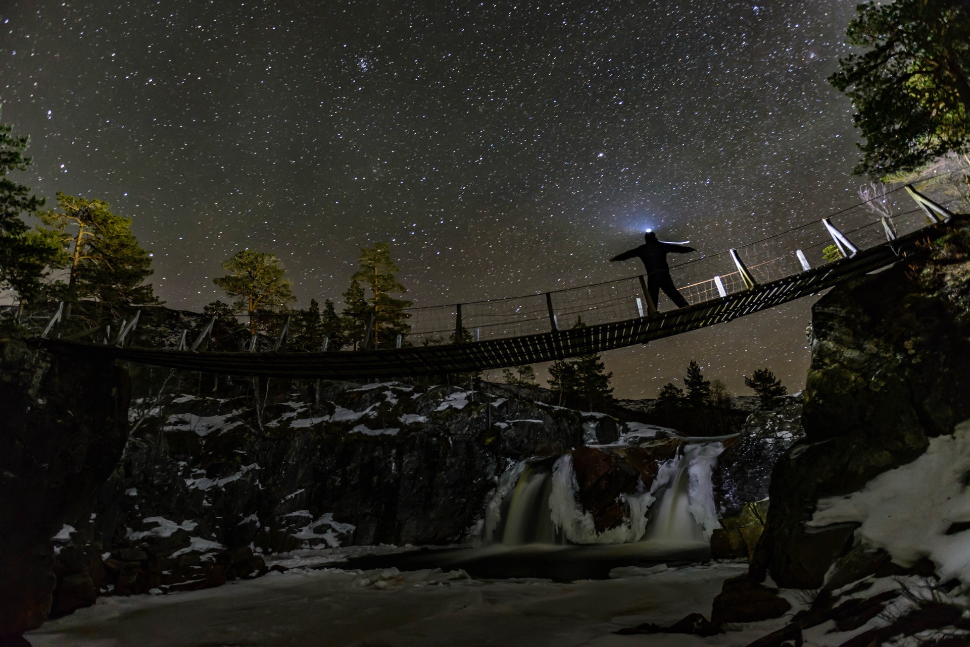 Man on suspension bridge under starry sky with waterfall HD Wallpaper