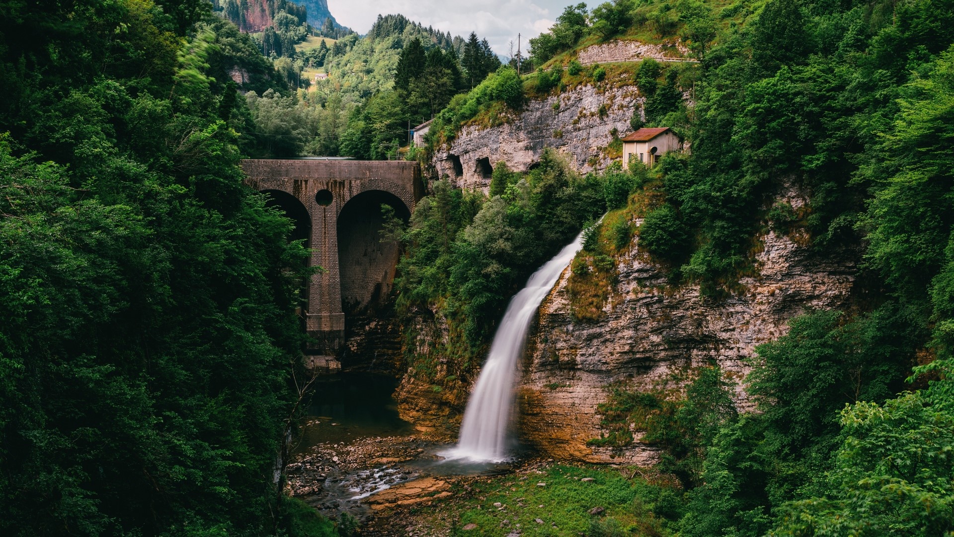 Waterfall cascading near a stone bridge in a green mountainous landscape HD Wallpaper
