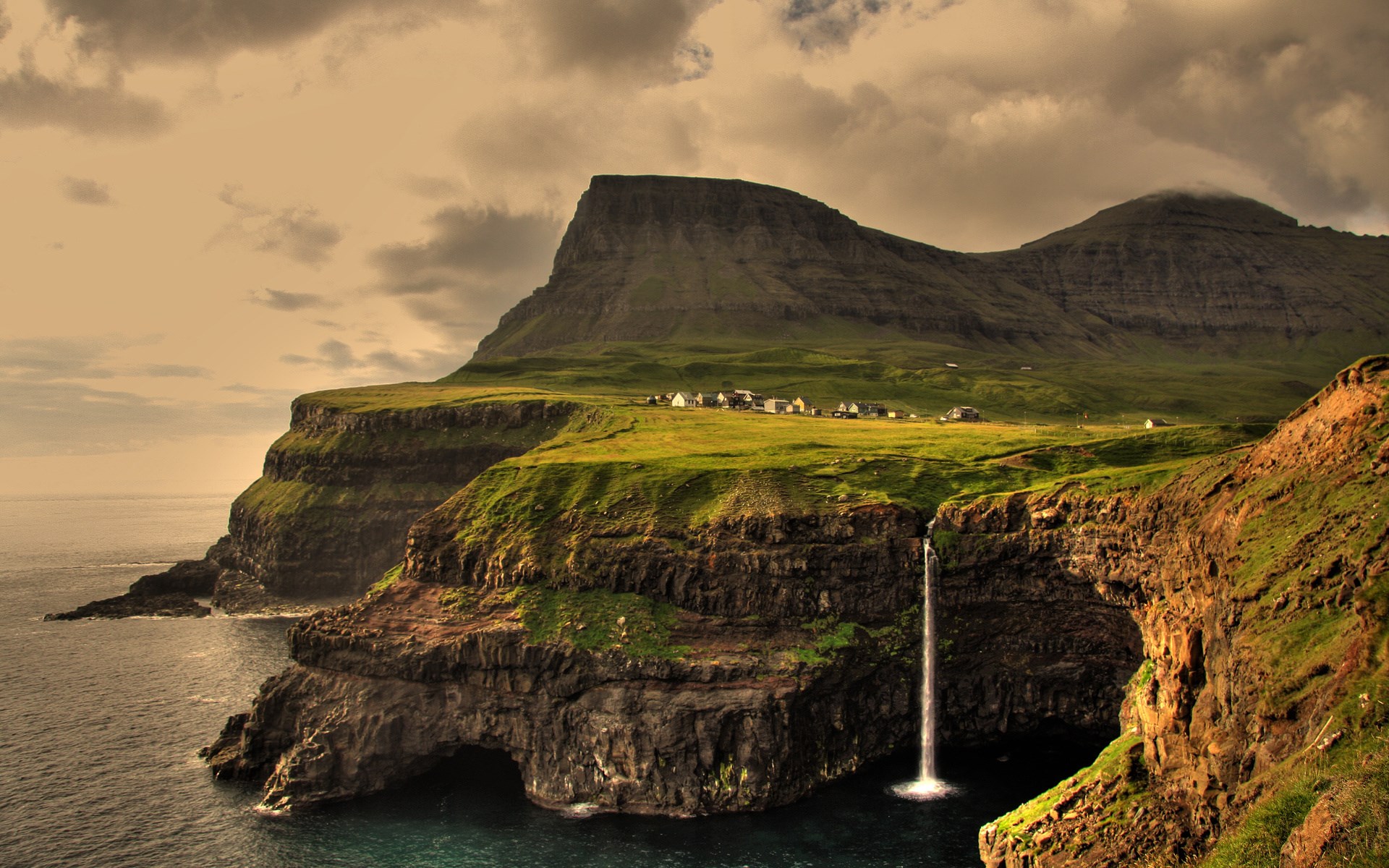 Waterfall cascading from cliffs into the sea near a village HD Wallpaper