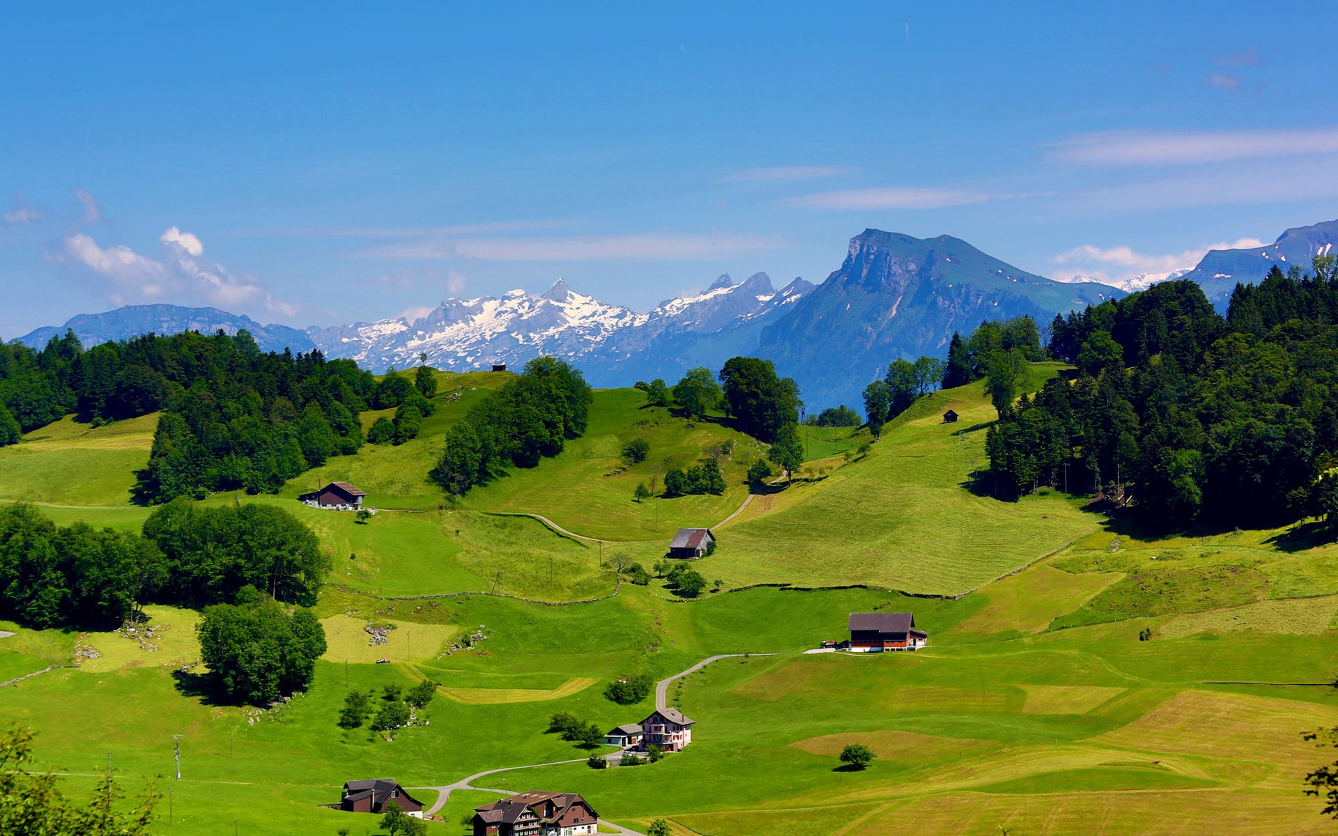 Green rolling hills with houses and snow-capped mountains HD Wallpaper