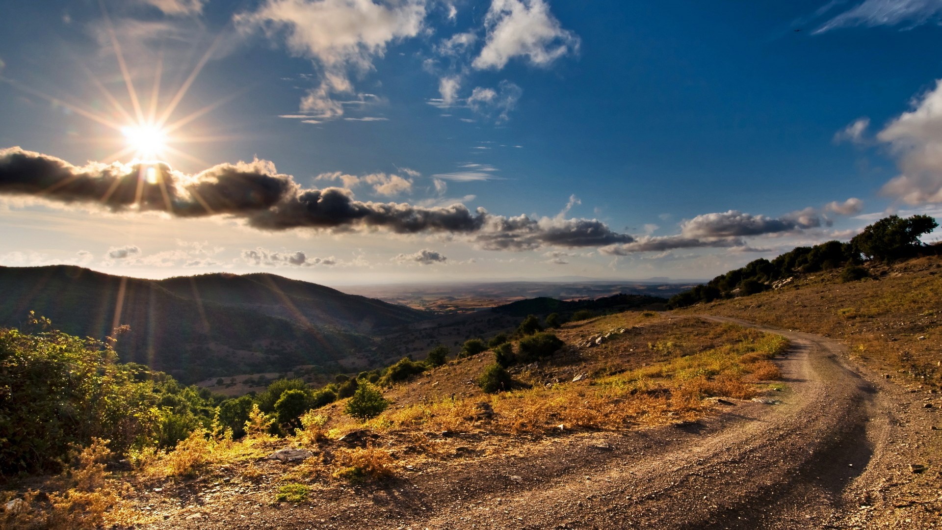 Sunbeams over a mountain landscape with a dirt road HD Wallpaper