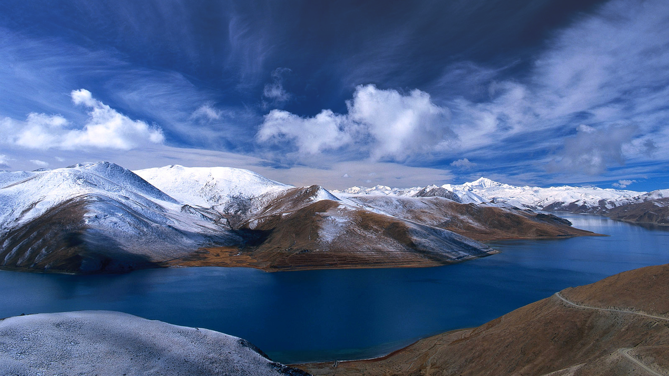 Snow-capped mountains surrounding a blue lake under a cloudy sky HD Wallpaper